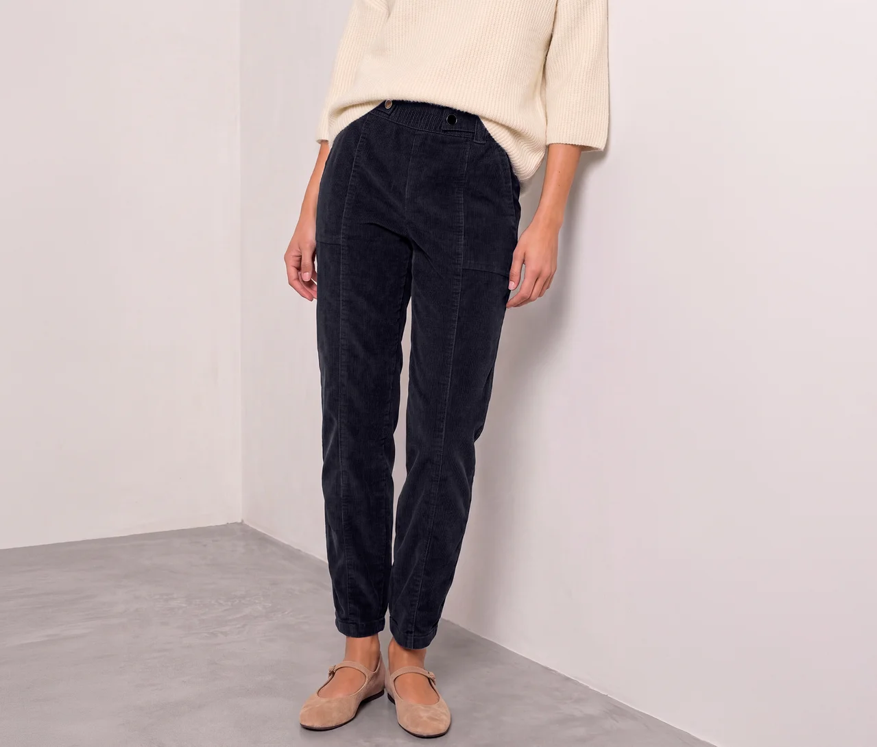Eine Person trägt einen cremefarbenen Pullover, eine dunkelblaue Street One Cordhose und hellbraune Ballerinas, stehend an einer weißen Wand.