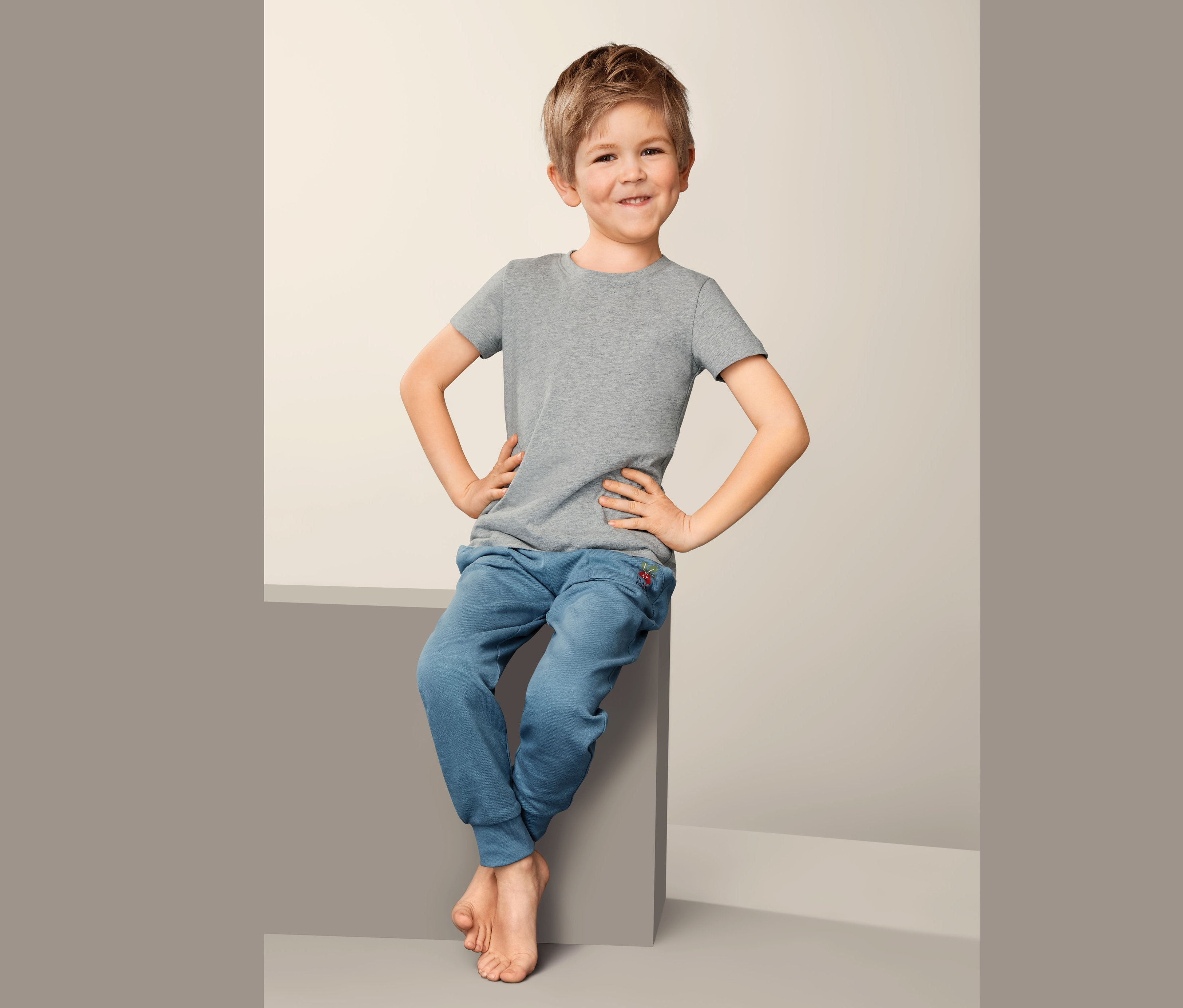 Ein Junge sitzt und trägt 2 Unterhemden und blaue Jogginghosen.