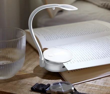 Auf einem Nachttisch liegen ein offenes Buch, eine LED-Leseleuchte, ein Glas Wasser und eine Armbanduhr.