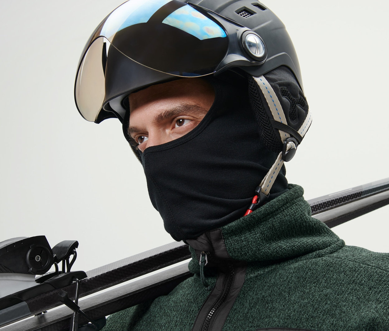 Mann mit Skihelm und Performance-Sturmhaube trägt Ski über der Schulter.