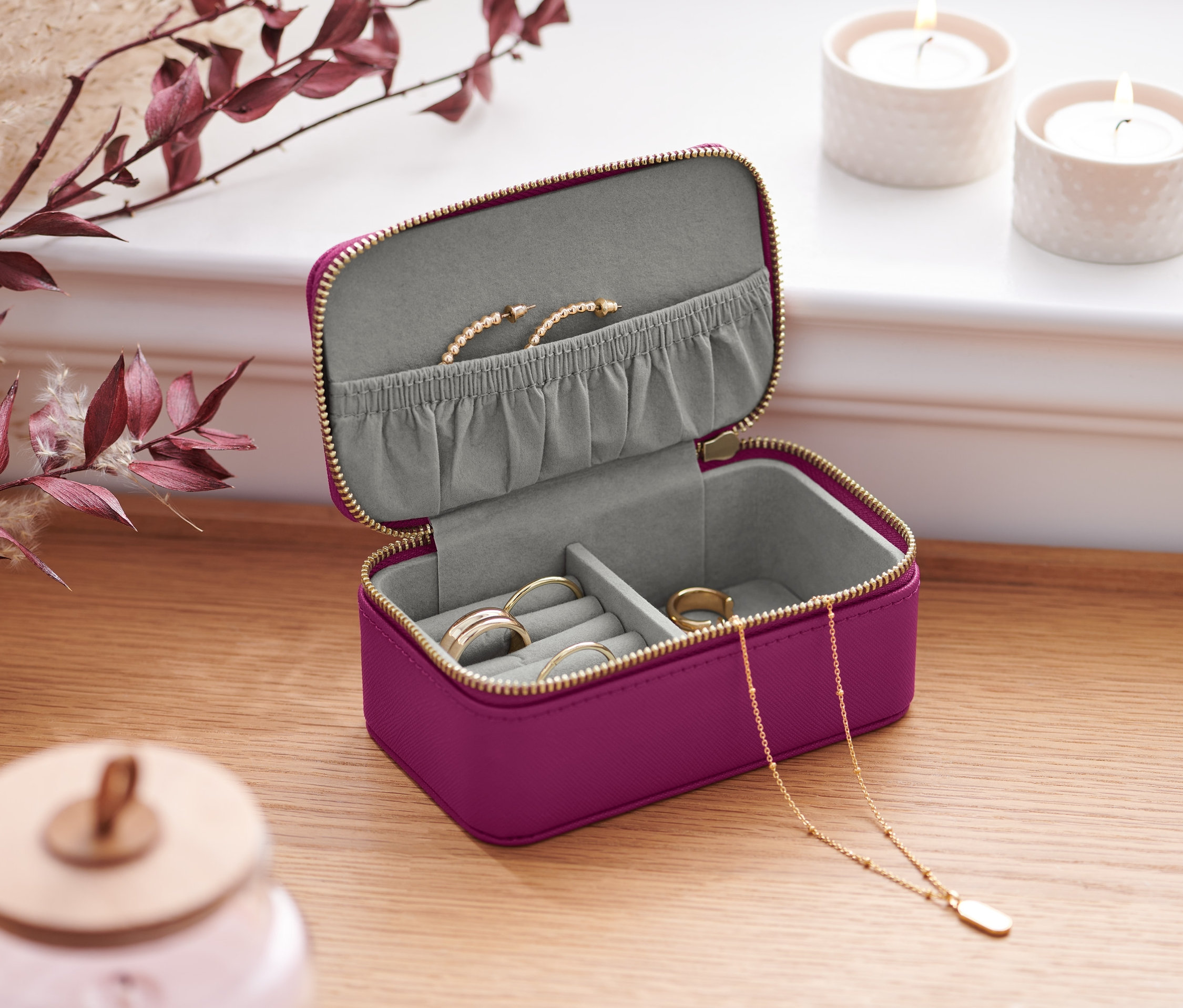 Geöffnete kleine Schmuckdose in Lila mit goldenem Reißverschluss auf einem Holztisch. Darin befinden sich Ringe, eine Halskette und ein Armband.