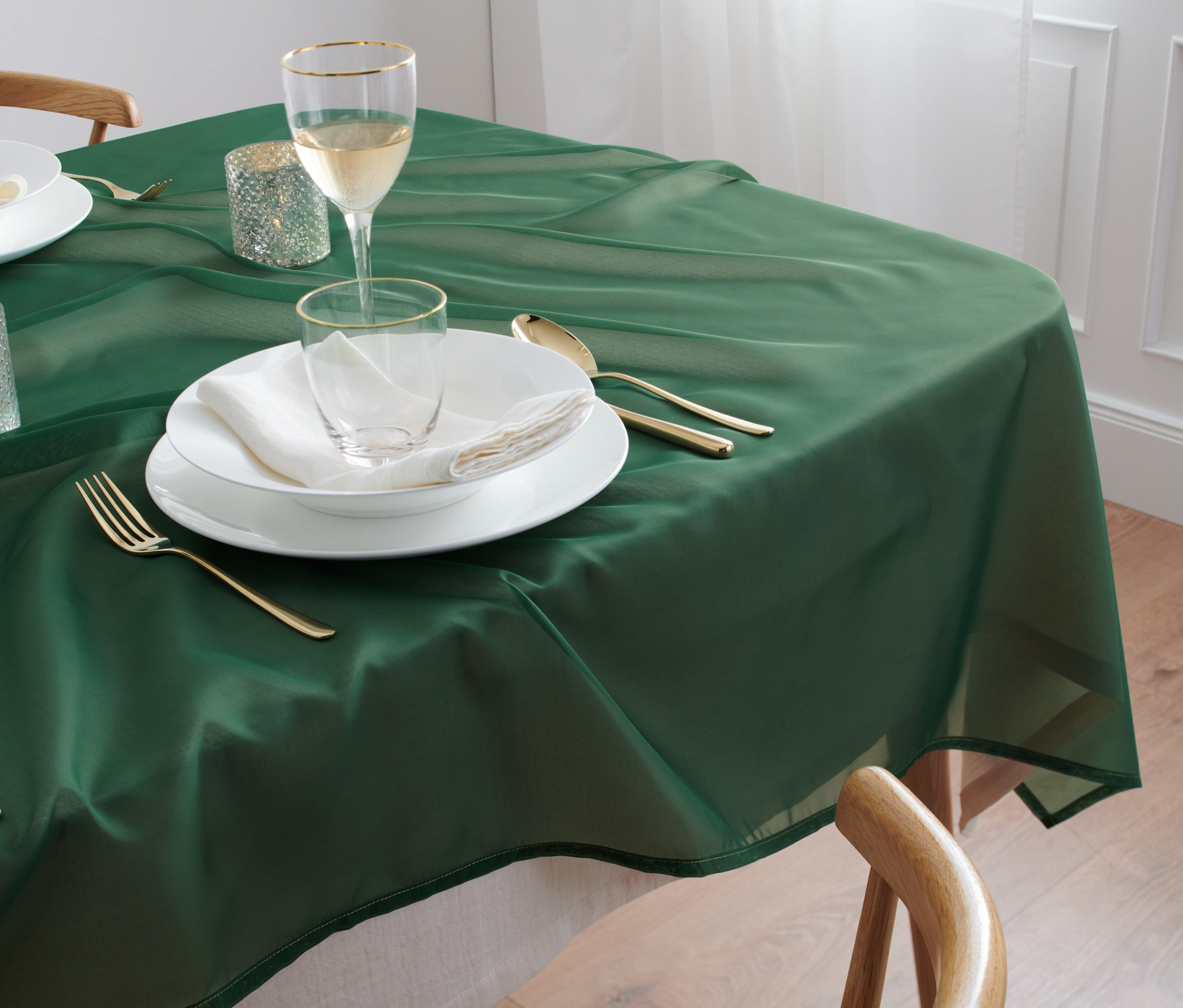 Eine grüne Organza-Tischdecke, ca. 140 x 180 cm, liegt auf einem Tisch mit Gedeck, das weiße Teller, Gläser und goldenes Besteck umfasst.