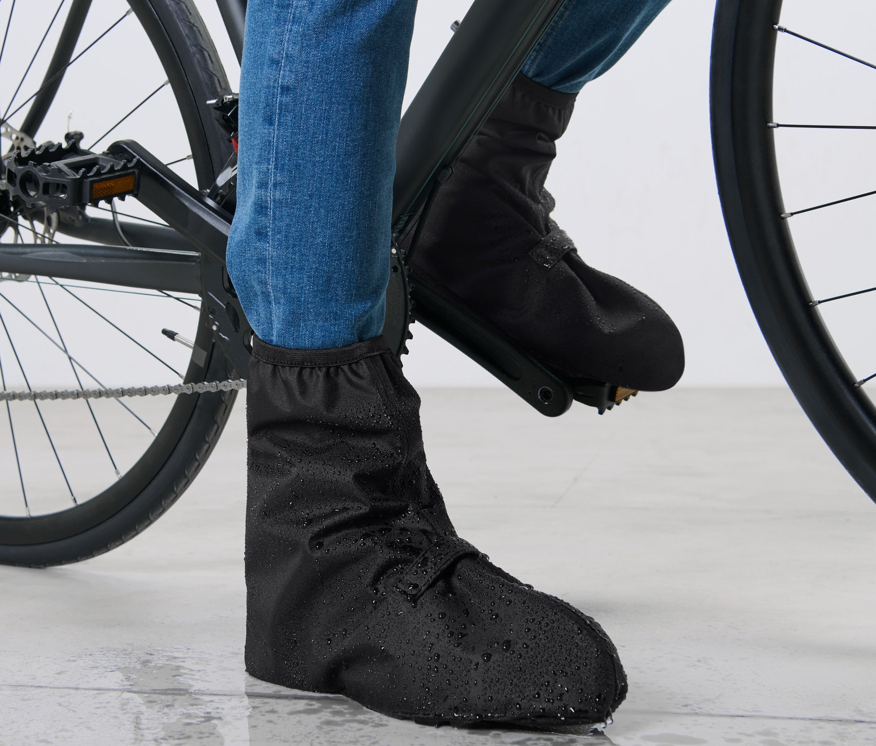 Füße eines Radfahrers in blauen Jeans und schwarzen Regen-Überschuhen auf einem Fahrrad.