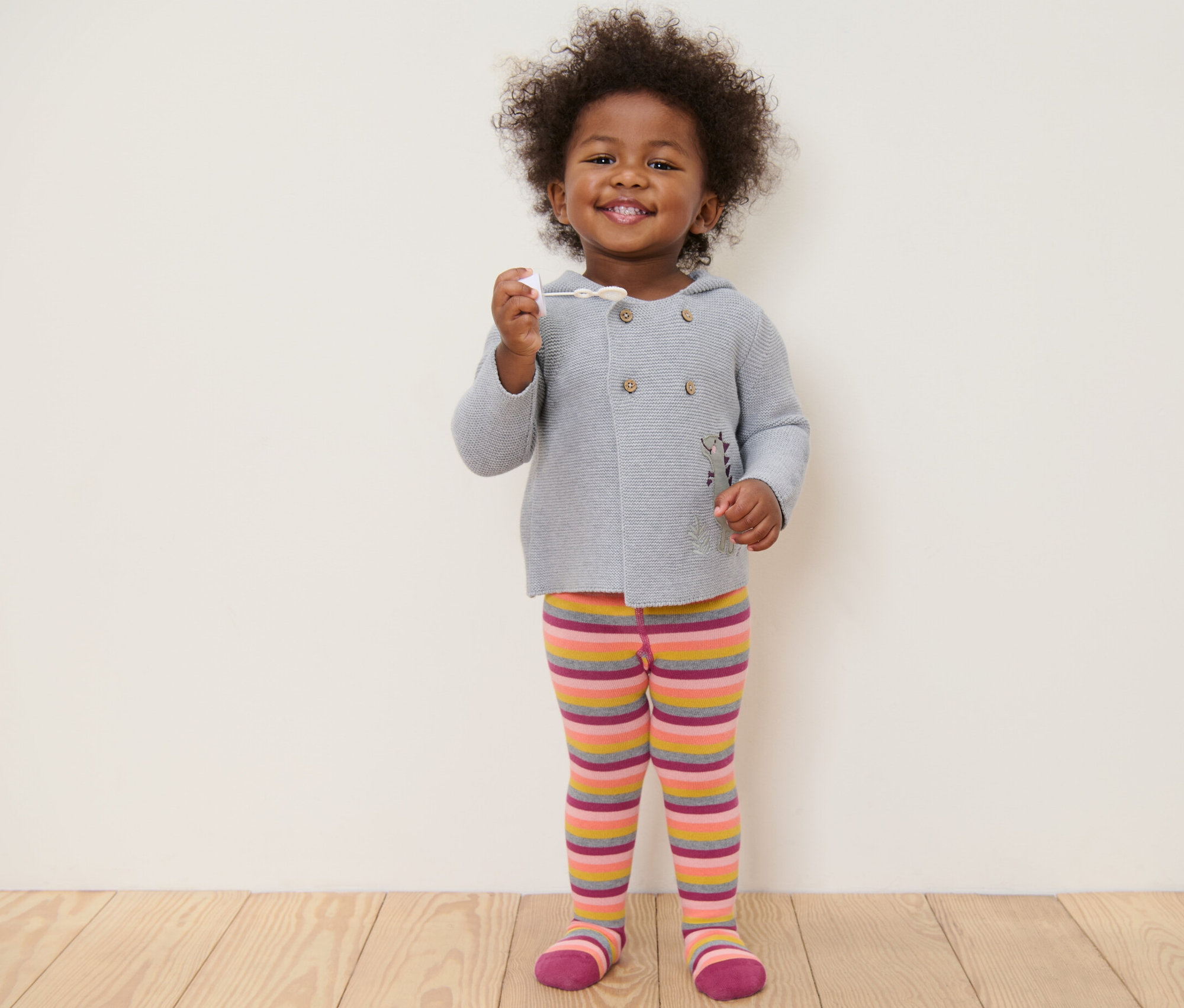 Ein lächelndes Kind steht und hält ein Spielzeug in der Hand. Es trägt einen grauen Pullover und rosa gestreifte Strumpfhosen.