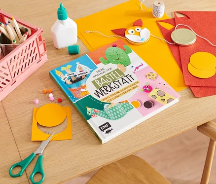Auf einem Tisch liegen das Buch "Meine nachhaltige Bastelwerkstatt", eine Schere, bunte Papiere, Kleber und Buntstifte.