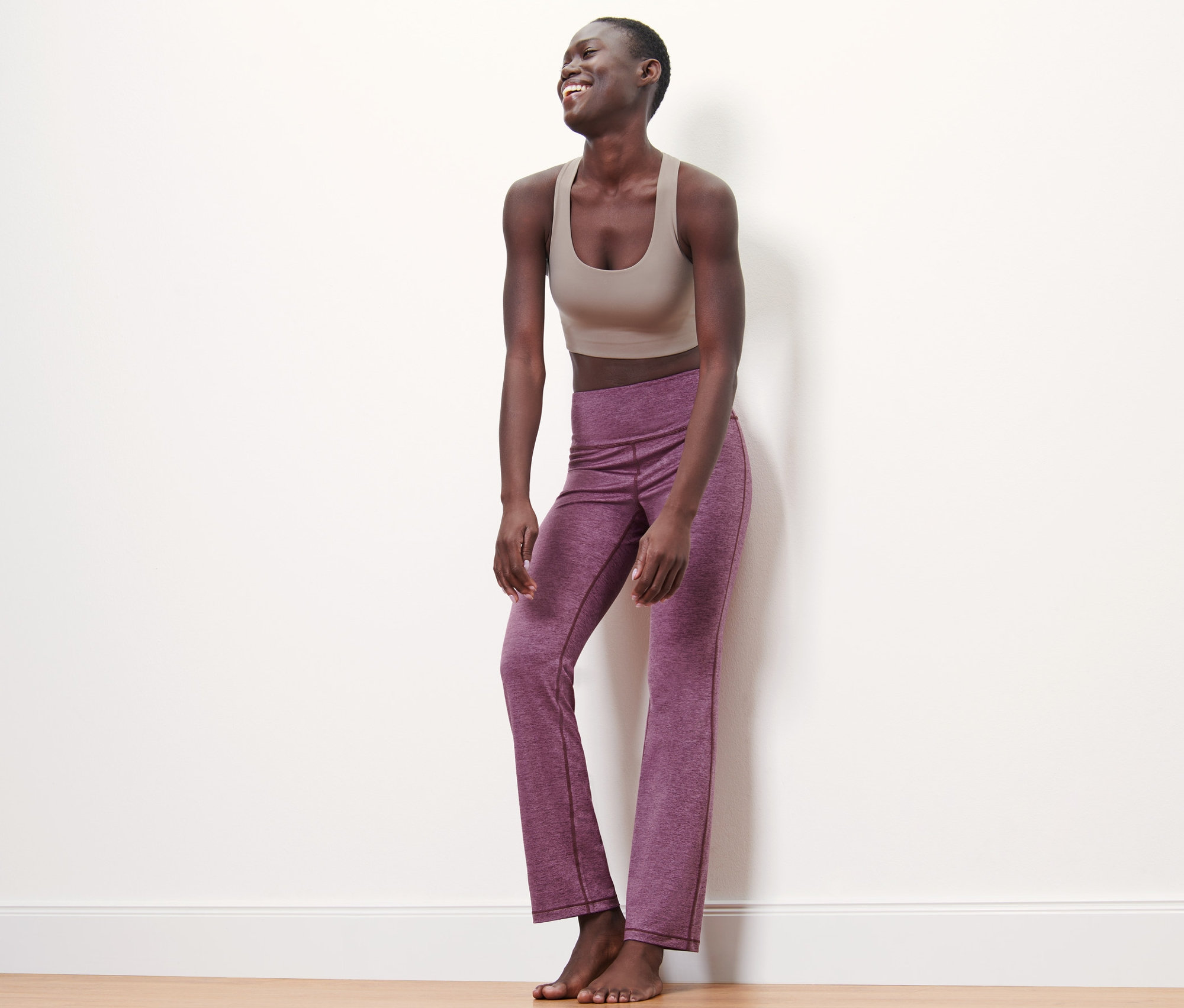 Eine Frau lacht und steht an einer Wand in einem braunen Sport-BH und einer Bodyforming-Sporthose in Mauve.