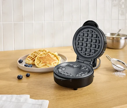 Offener, schwarzer Waffelautomat neben einem Teller mit Waffeln und Blaubeeren auf einem Holztisch.
