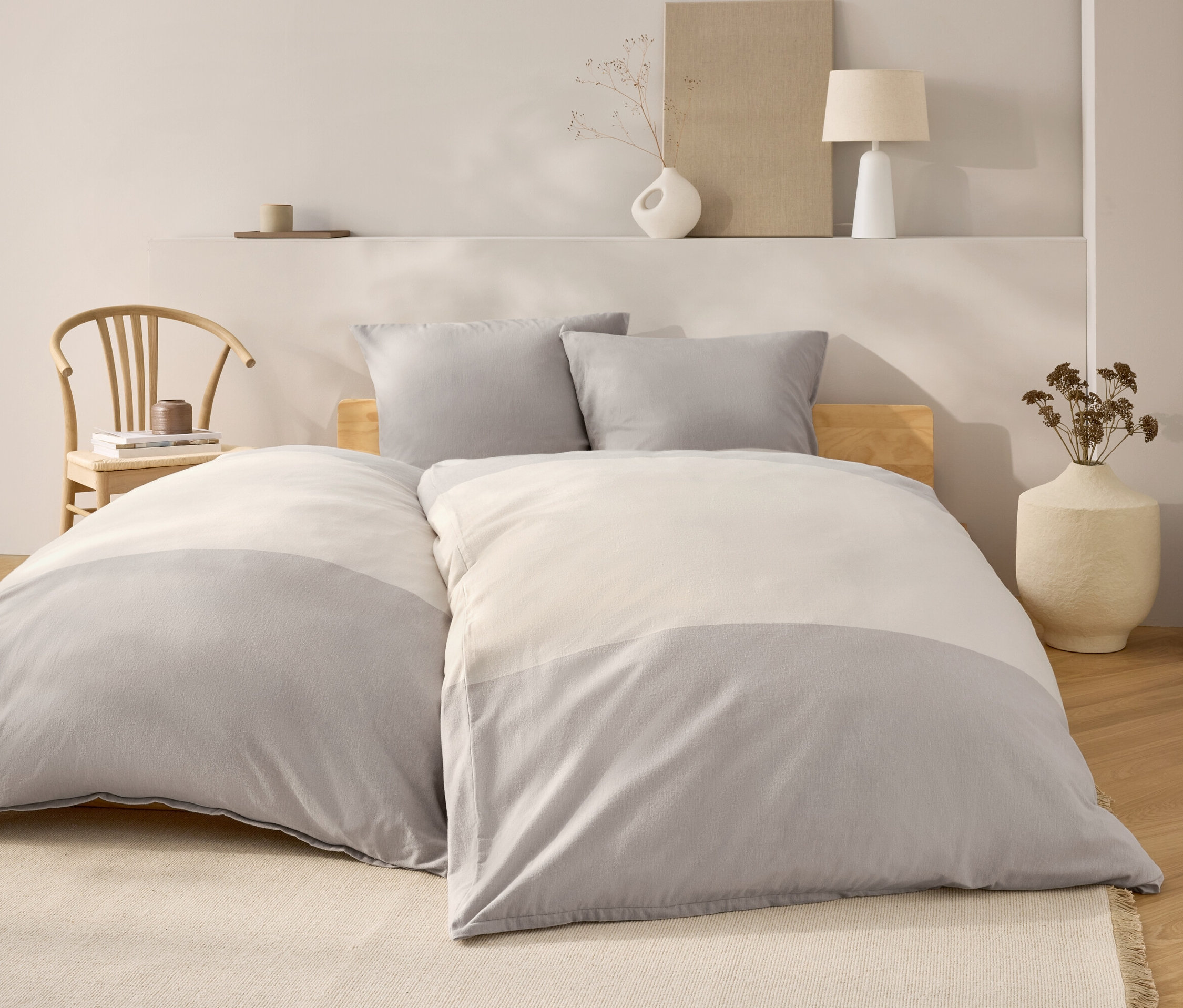 Schlafzimmer mit Bett mit grau-weisser Bettwäsche, zwei grauen Kissen und einem hölzernen Kopfteil. Im Hintergrund ein Holzstuhl und Dekoration.