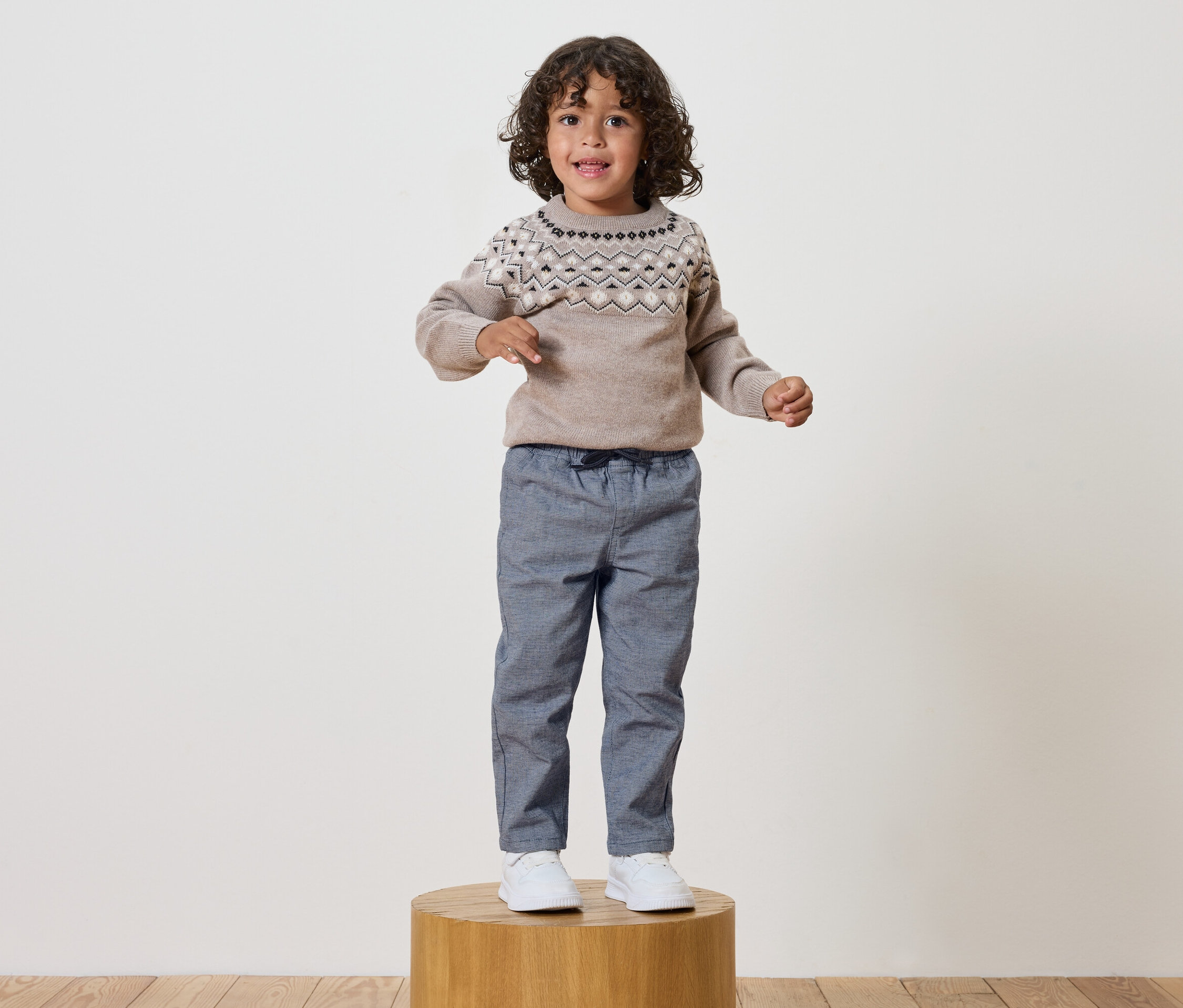 Junge mit lockigem Haar steht auf einem Holzsockel und trägt einen gemusterten Pullover und blaue Hosen.