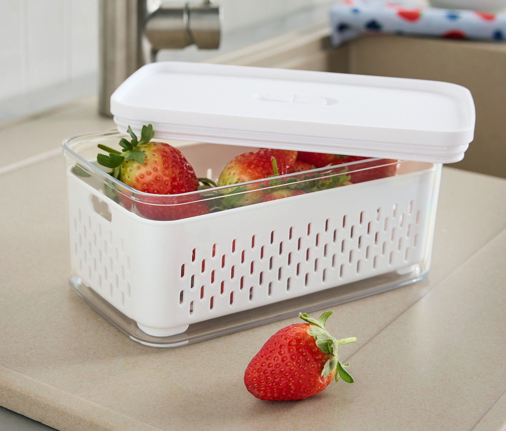 Eine teilweise geöffnete Frischhaltedose mit Siebeinsatz, gefüllt mit Erdbeeren, steht auf einer Küchentheke neben einem Wasserhahn.