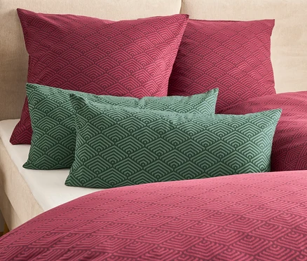Auf einem Bett liegen zwei weinrote und zwei grüne Kissen mit geometrischem Muster.