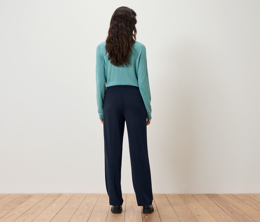 Rückansicht einer Frau mit dunkelbraunem Haar, die einen türkisfarbenen Pullover und dunkelblaue Hosen trägt.
