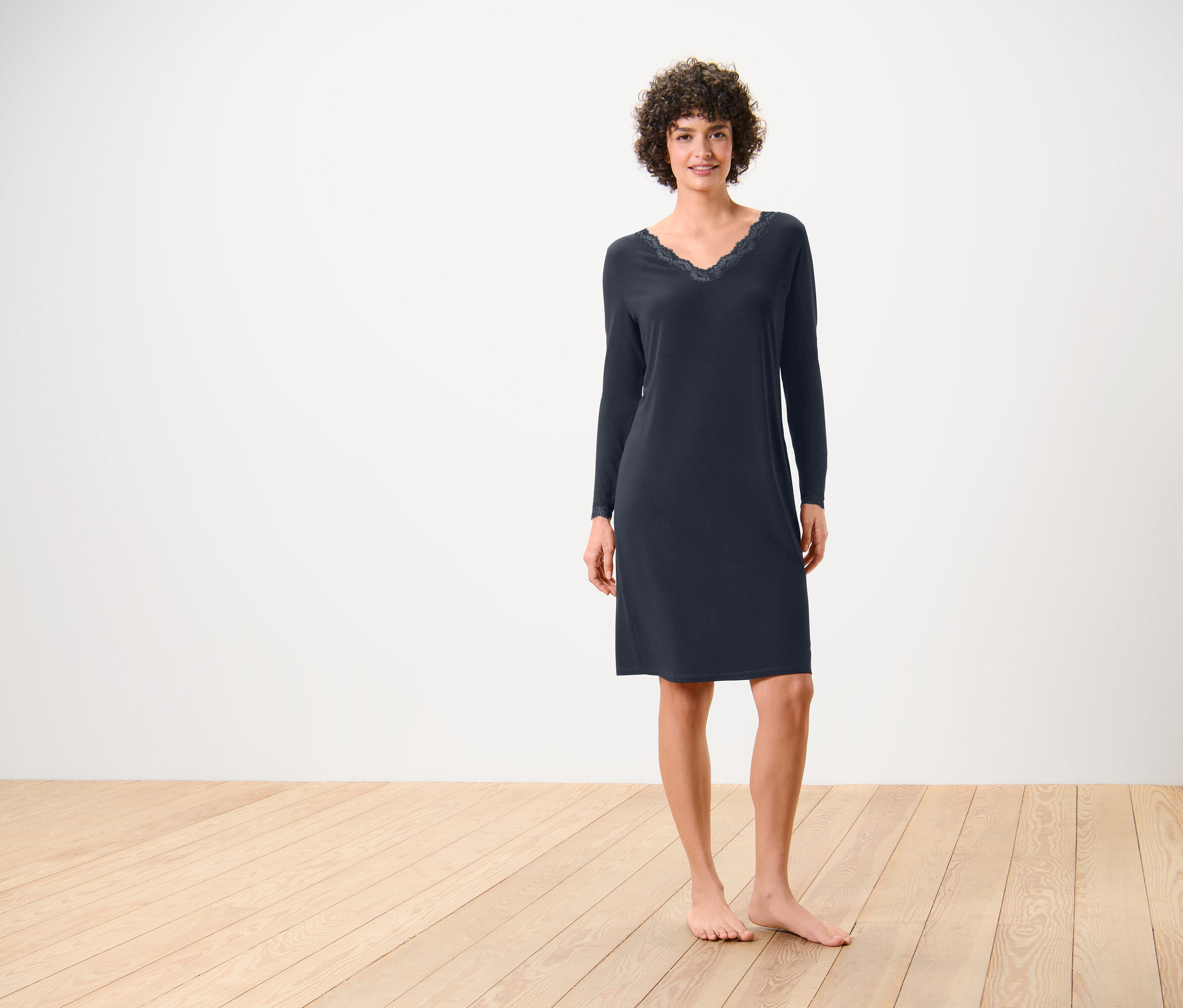 Frau steht barfuß auf Holzboden, sie trägt ein nachtblaues, langärmeliges Nachthemd mit Spitze am Ausschnitt.