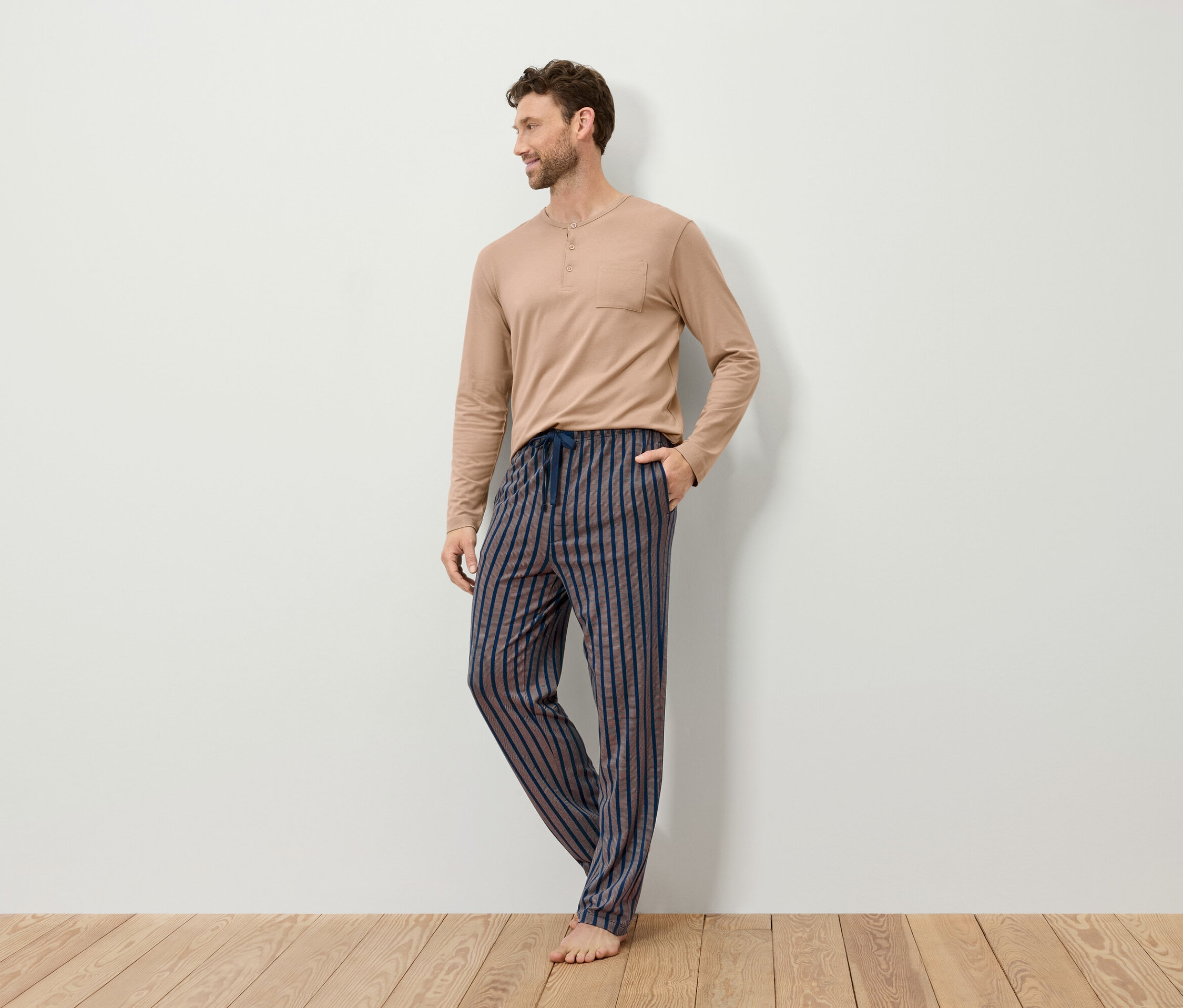 Mann trägt ein beiges, langärmeliges Schlafanzugoberteil und eine gestreifte Schlafanzughose.