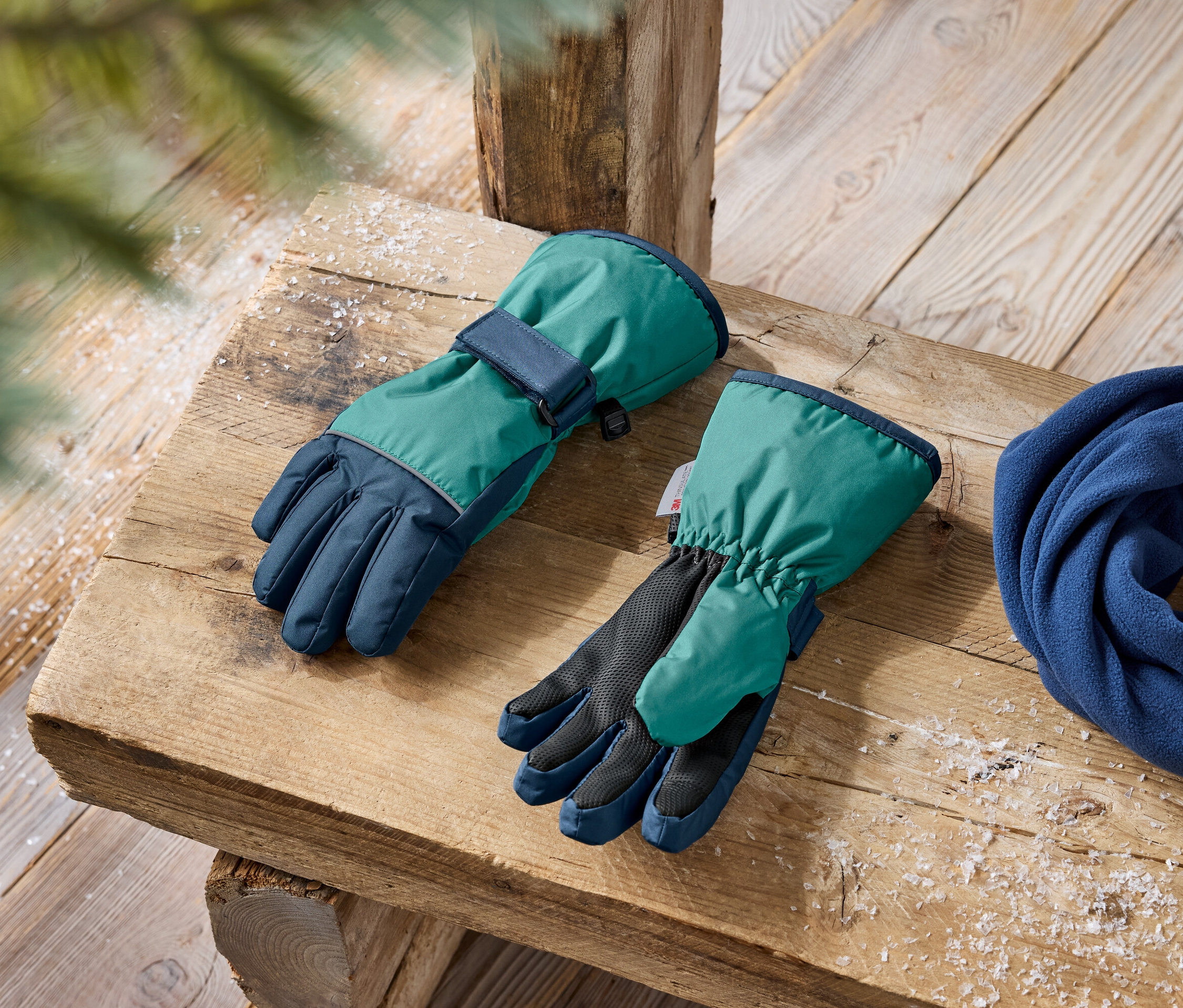 Winterhandschuhe in Grün und Dunkelblau und ein blauer Fleece-Schal liegen auf einer Holzbank.