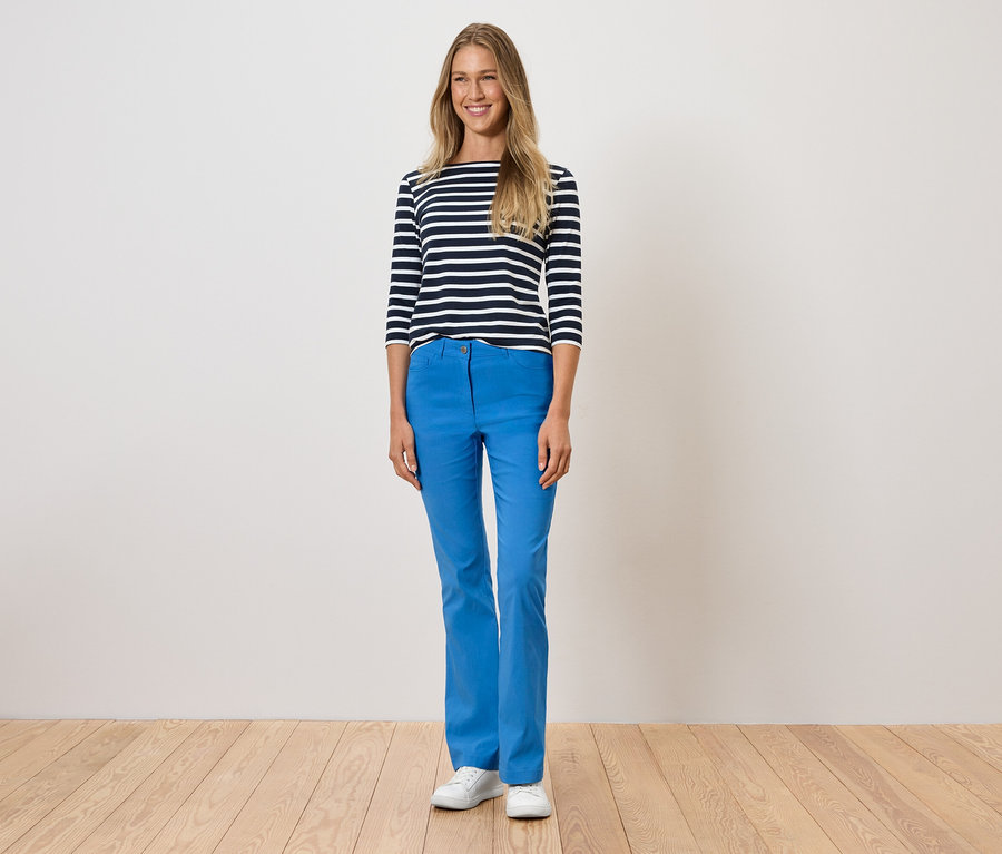 Frau steht und trägt blaue Hosen, ein gestreiftes T-Shirt und weiße Schuhe.