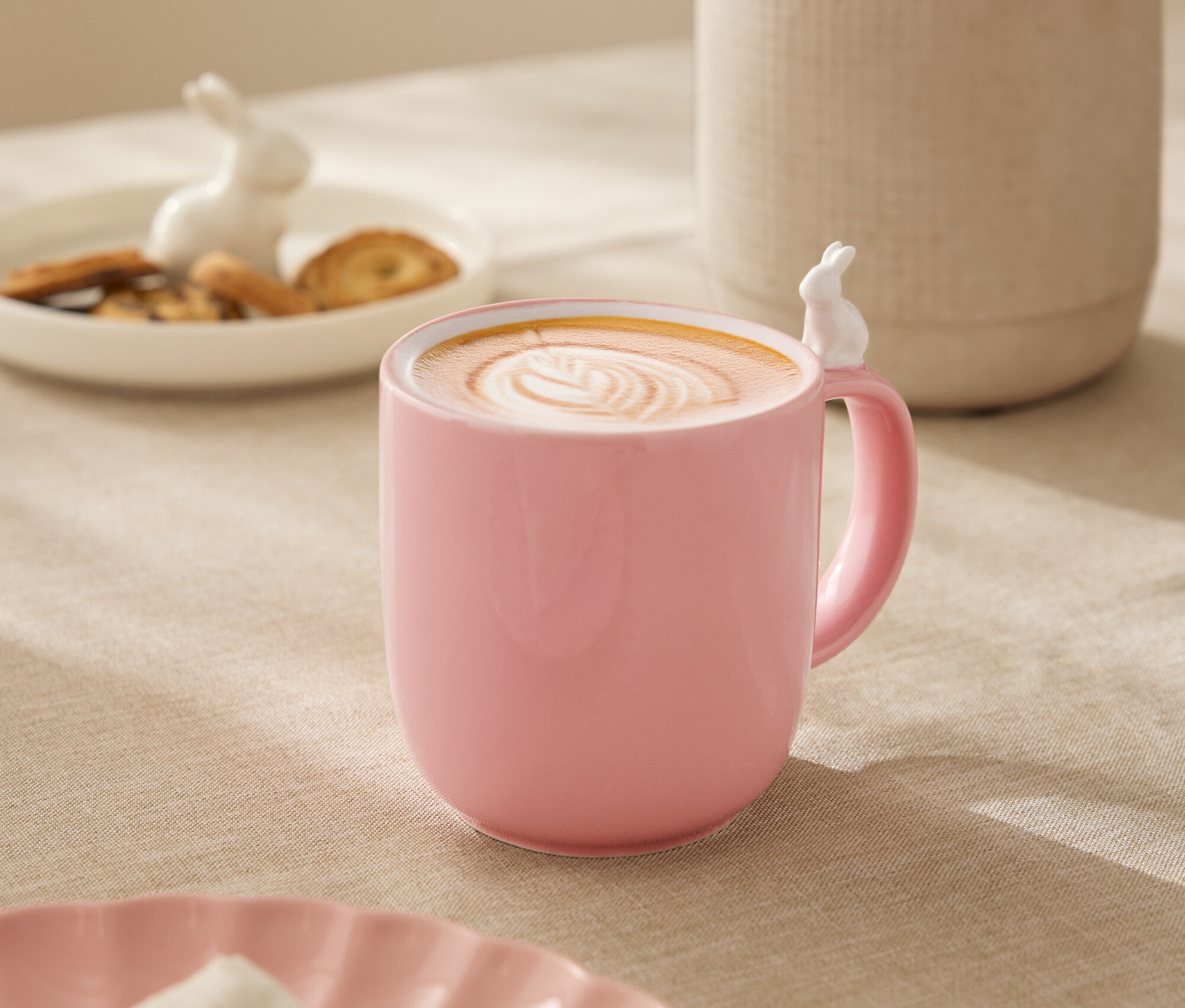 Auf einem Tisch steht eine rosa Tasse mit einem weißen Hasen am Rand, gefüllt mit einem Getränk mit Latte Art. Im Hintergrund befinden sich eine weiße Vase mit Blumen, ein Teller mit Keksen und eine Hasenfigur.