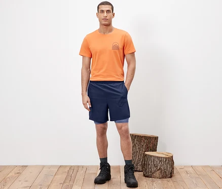 Mann posiert in 2-in-1-Funktionsshorts, orangefarbenem T-Shirt und Wanderstiefeln.