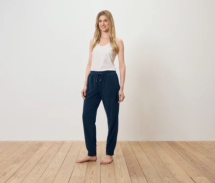 Eine Frau steht auf einem Holzboden und trägt ein weißes Top und eine dunkelblaue Trainingshose.