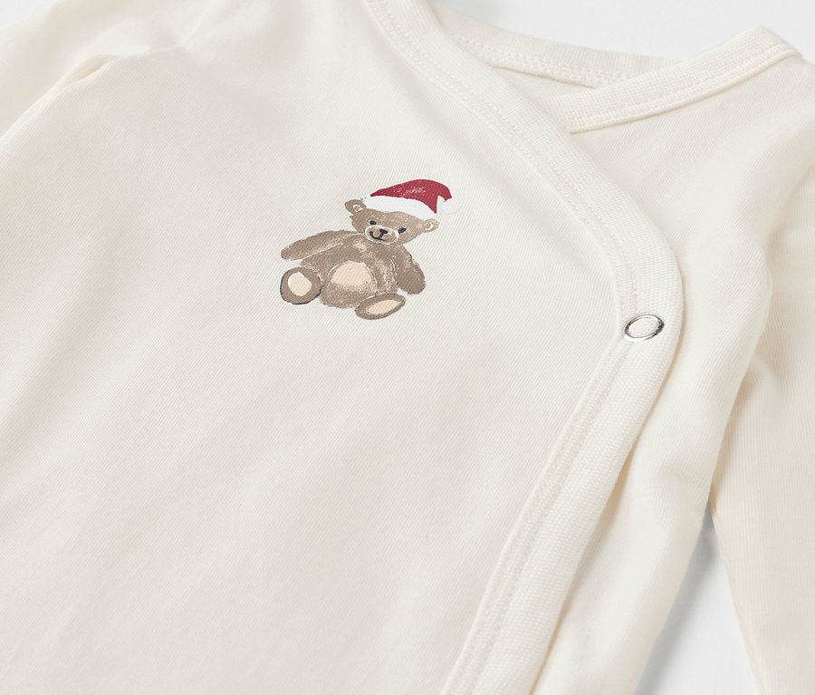 Detailaufnahme eines weißen Babybodys mit einem braunen Teddybär mit roter Mütze.