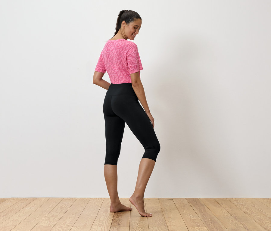 Frau in rosa T-Shirt und schwarzen Leggings steht auf einem Holzboden.