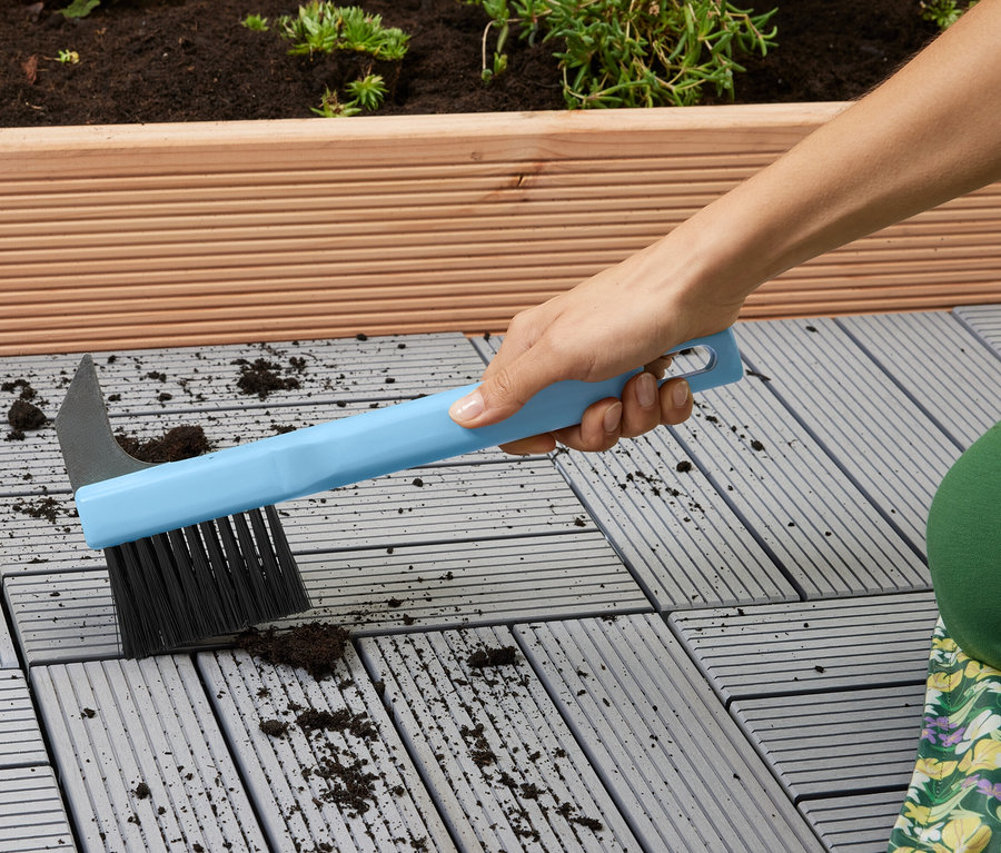 Frau reinigt graue Terrassenplatten mit einer blauen Bürste.