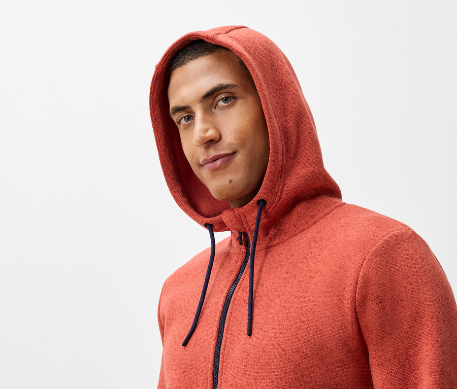 Porträt eines Mannes mit Kapuze und dunkelblauem Reißverschluss auf einem orangefarbenen Sweatshirt.
