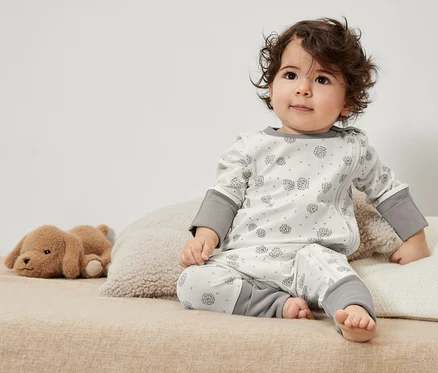 Ein lockiges Kleinkind sitzt in 3 Baby-Pyjamas mit einem Spielzeug auf dem Bett.