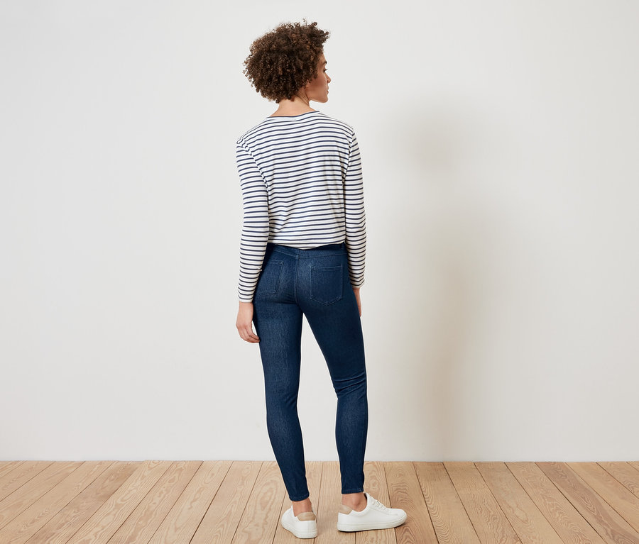 Frau steht mit dem Rücken zum Betrachter, trägt ein gestreiftes Oberteil, dunkle Jeans und weiße Turnschuhe.