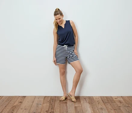 Frau posiert vor einer weißen Wand, trägt ein marineblaues Top und gestreifte Shorts.