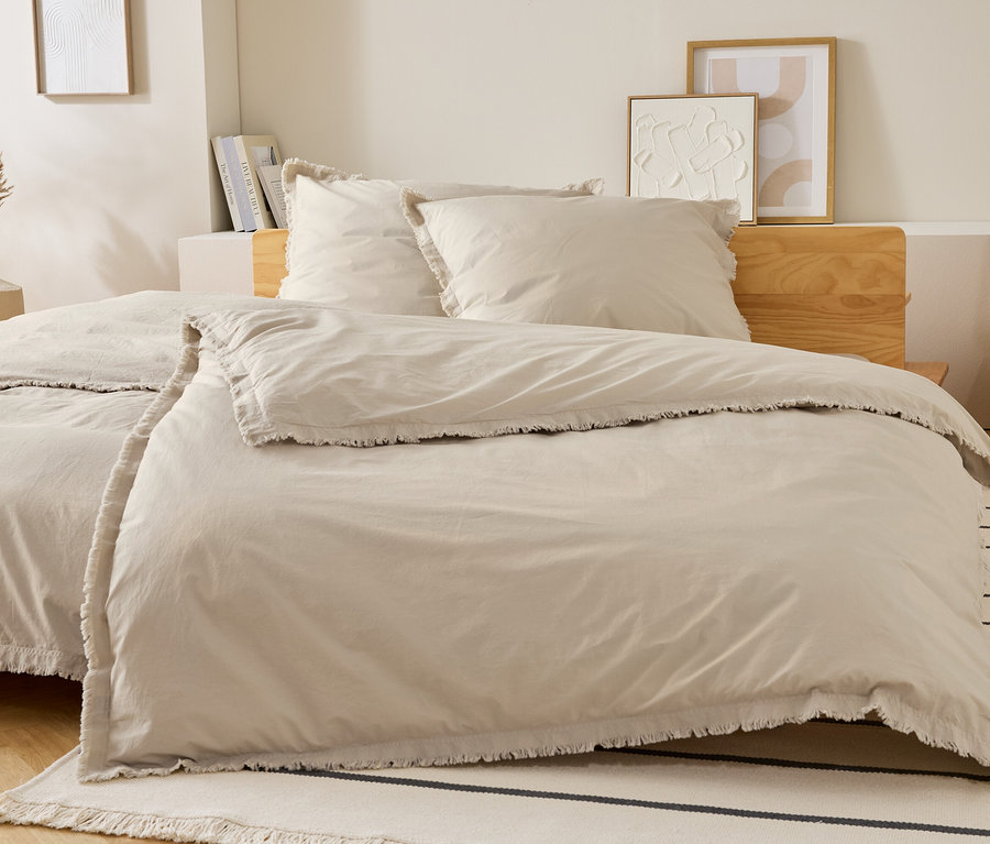 Beige Bettwäsche mit Fransen liegt auf einem Bett mit zwei Kissen. Dekorationen und ein Nachttisch stehen im Hintergrund.