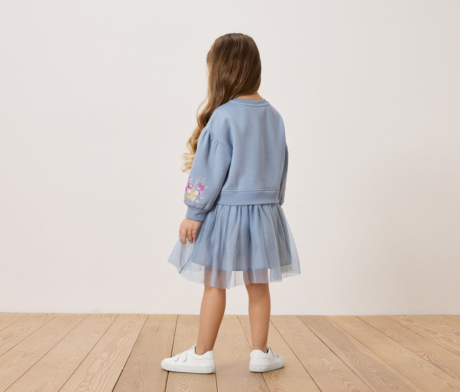 Rückansicht eines kleinen Mädchens mit langen braunen Haaren, das ein blaues Sweatshirt mit Blumenmuster auf dem Ärmel und einen passenden Tüllrock sowie weiße Turnschuhe trägt.
