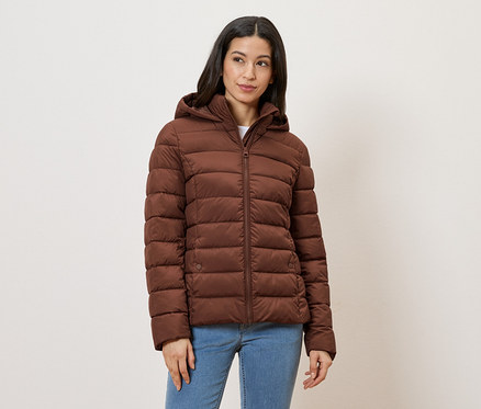 Frau mit Jeans und brauner Steppjacke mit Kapuze vor hellem Hintergrund.