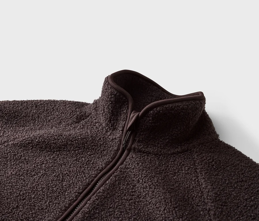 Nahaufnahme einer dunkelbraunen Fleecejacke mit Reißverschluss und hohem Kragen vor einem weißen Hintergrund.
