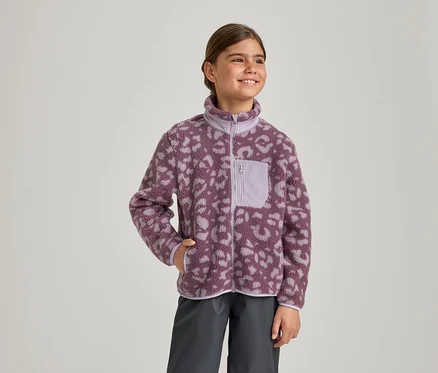 Ein junges Mädchen lächelt und blickt nach rechts. Sie trägt eine lila-helllila Fleecejacke mit Leopardenmuster, Reißverschluss und Brusttasche sowie dunkelgraue Hosen. Ihre linke Hand steckt in der Jackentasche.