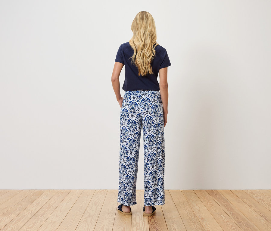 Rückansicht einer blonden Frau mit marineblauem T-Shirt und blau-weiß gemusterter Hose.