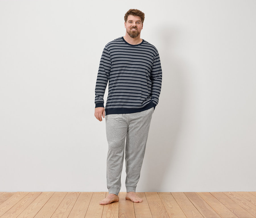 Mann steht barfuß vor einer Wand und trägt einen grauen Pyjama.