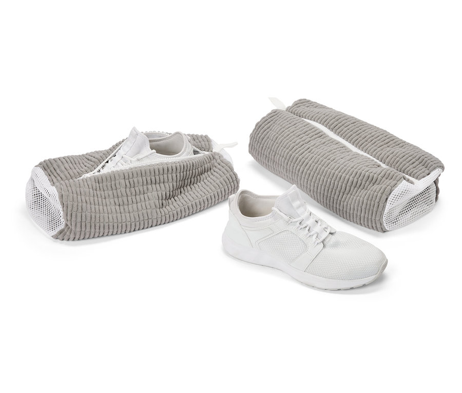Ein weißer Turnschuh vor einem grauen Schuhbeutel. Ein weiterer Schuh steckt in einem offenen Beutel, ein dritter Beutel ist geschlossen.
