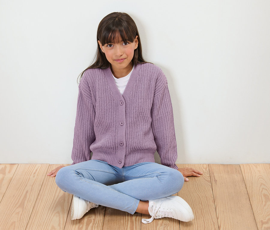Mädchen sitzt im Schneidersitz auf einem Holzboden, trägt einen lila Strickjacke, blaue Jeans und weiße Turnschuhe.