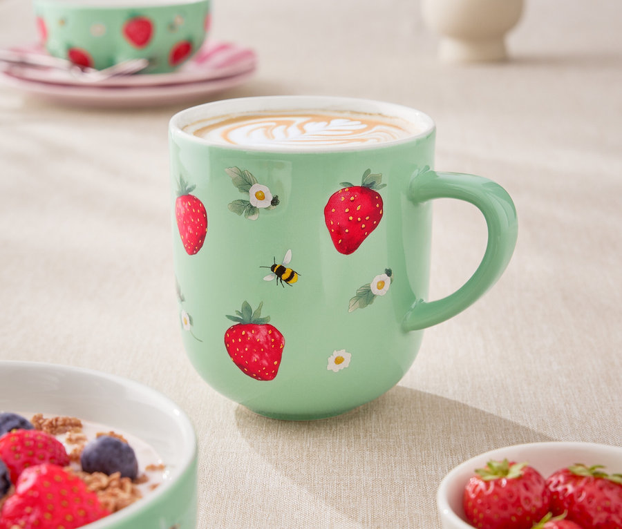 Nahaufnahme eines Tisches mit einer hellgrünen Tasse Kaffee, einer Müslischale mit Joghurt und Beeren sowie einer kleinen Schale mit Erdbeeren. Die Tasse ist mit Erdbeeren und kleinen Blumen verziert.