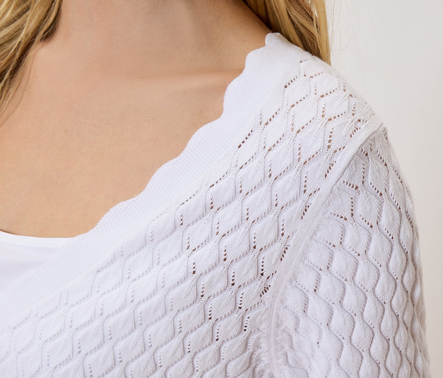 Nahaufnahme einer Frau mit blonden Haaren, die eine weiße Strickjacke mit Lochmuster über einem weißen Top trägt.