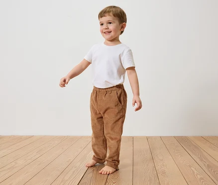 Ein lächelnder Junge mit braunen Haaren steht barfuß auf einem Holzboden und trägt ein weißes T-Shirt und braune Cordhosen.