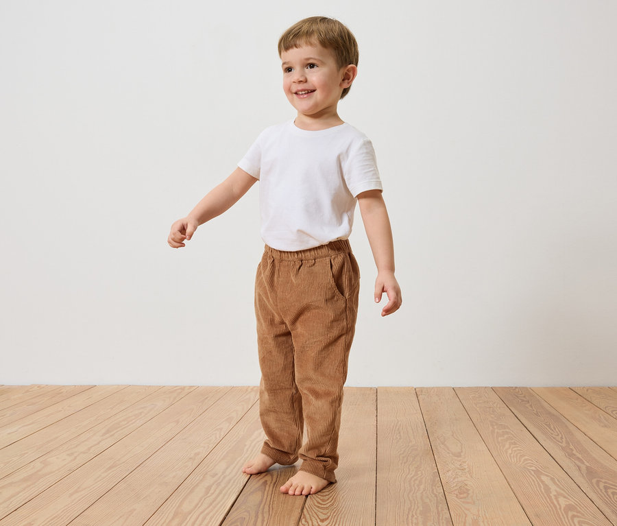 Ein lächelnder Junge mit braunen Haaren steht barfuß auf einem Holzboden und trägt ein weißes T-Shirt und braune Cordhosen.