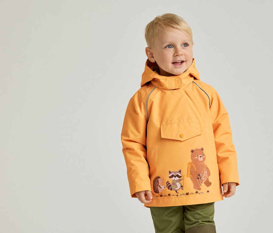 Kleiner Junge mit blonder Kurzhaarfrisur trägt eine orangefarbene Jacke mit Bären-, Waschbär- und Igelmotiv und grüne Hosen.