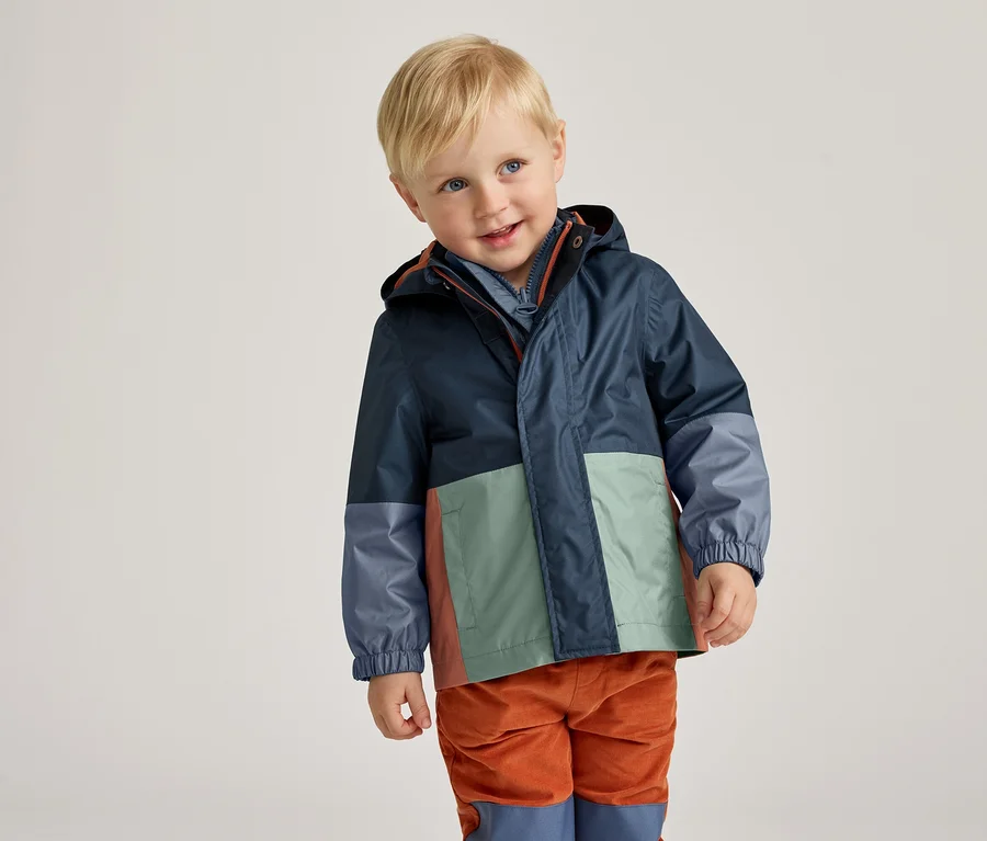 Ein lächelnder Junge mit blonden Haaren trägt eine marineblaue Regenjacke mit Farbblockdesign und orangefarbene Cordhosen.