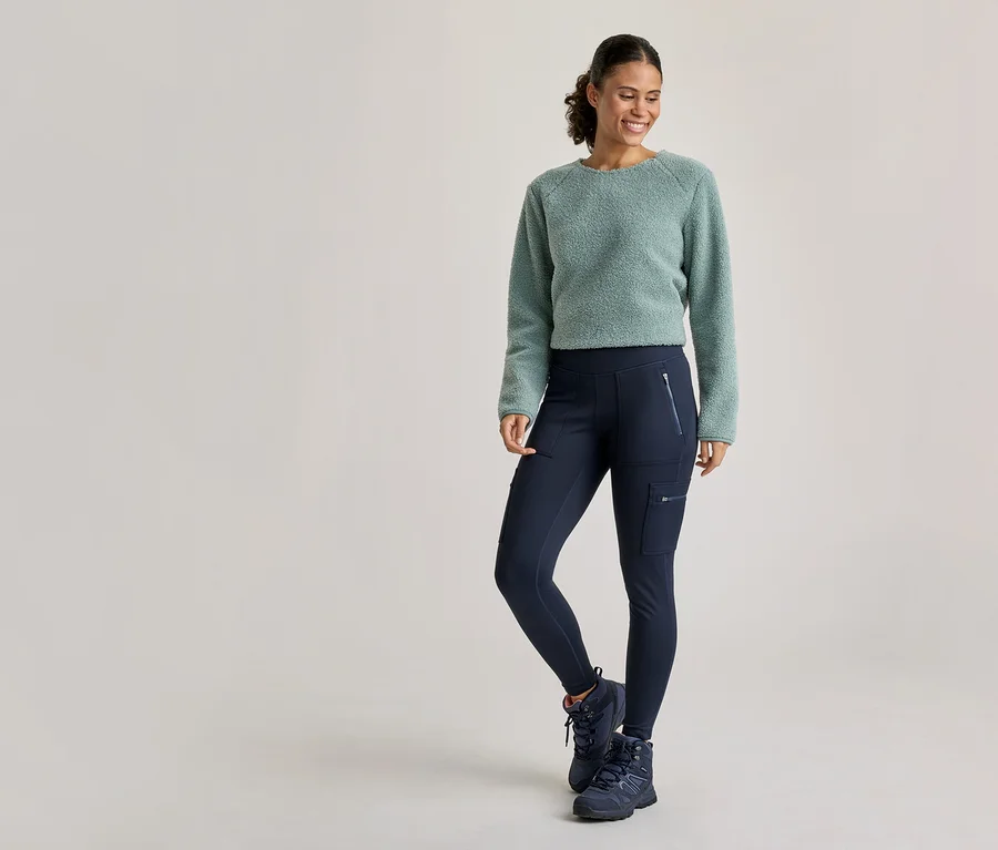 Eine lächelnde Frau trägt einen hellgrünen flauschigen Pullover, dunkelblaue Cargoleggings und dunkelblaue Wanderschuhe und steht mit einem angewinkelten Bein da.