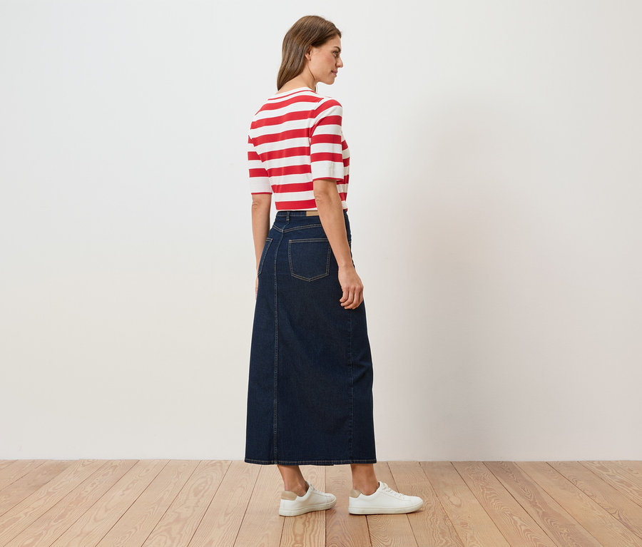 Frau von hinten, trägt ein rot-weiß gestreiftes Oberteil, einen langen Jeansrock und weiße Turnschuhe.