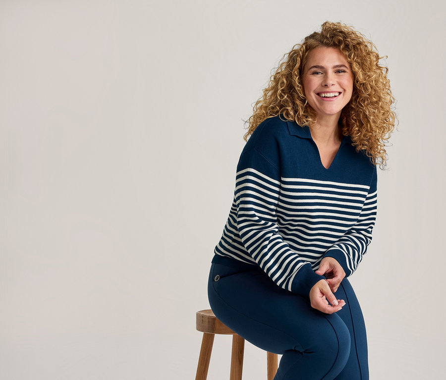 Lachende Frau mit Locken, die einen marineblauen und weiß gestreiften Pullover und marineblaue Hosen trägt, sitzt auf einem Holzschemel.