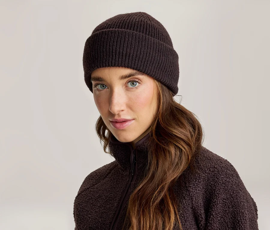 Frau mit langen braunen Haaren, die eine dunkelbraune gerippte Strickmütze und eine dunkelbraune Fleecejacke trägt und in die Kamera blickt.