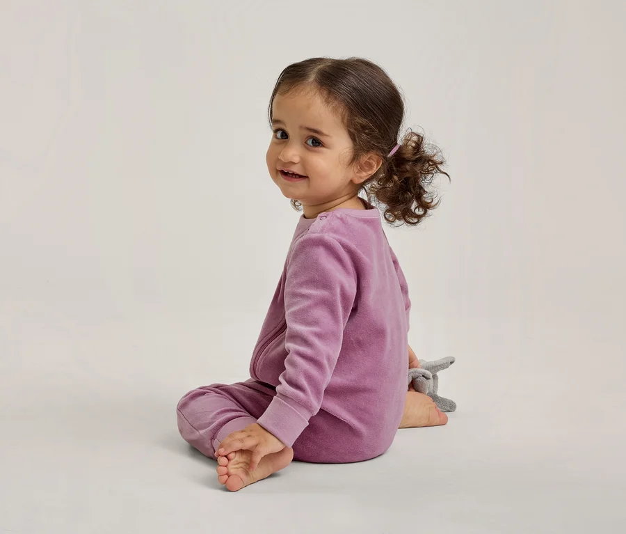 Ein Kleinkind mit dunklen Locken, bekleidet mit einem lila Samt-Langarmshirt und passender Hose, sitzt auf einem hellen Boden. Es blickt über seine linke Schulter und lächelt in die Kamera, mit einem kleinen grauen Stofftier daneben.